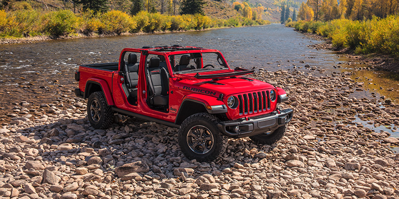 A red Jeep Gladiator with its doors off, sitting in a wilderness setting backed by a creek and woodlands.