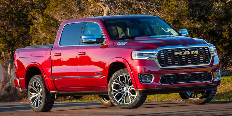 A red RAM 1500 sits on a parking pad backed by a green field and trees. 